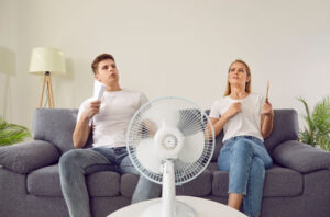 A man and woman sitting on a couch fanning themselves with a standing fan in front of them.