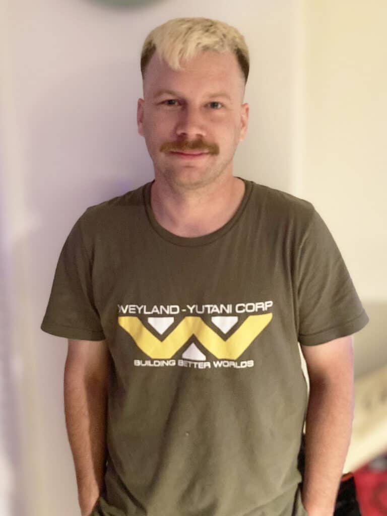 A man with light hair and a mustache stands indoors, wearing a Weyland-Yutani Corp t-shirt with "Building Better Worlds" text.