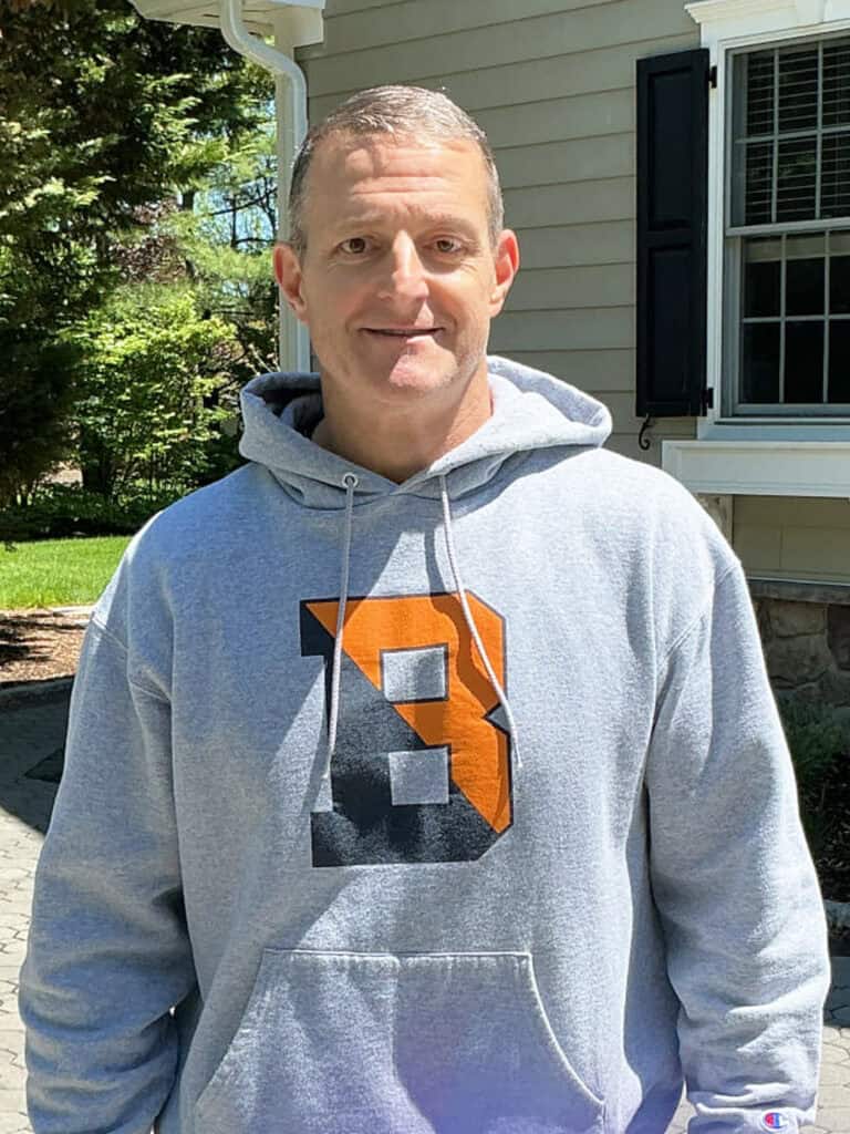 A man stands outdoors wearing a gray hoodie with a large letter "B" in black and orange on the front, in front of a house and greenery.