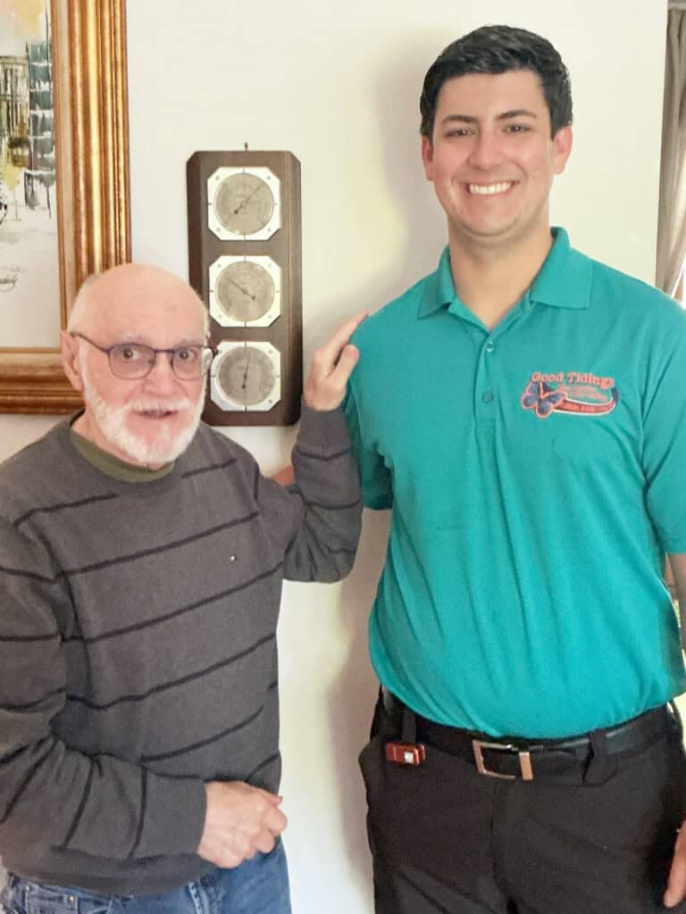 Two men stand indoors next to a wall-mounted weather station. The older man wears glasses and a striped sweater; the younger man wears a teal polo shirt and is smiling.