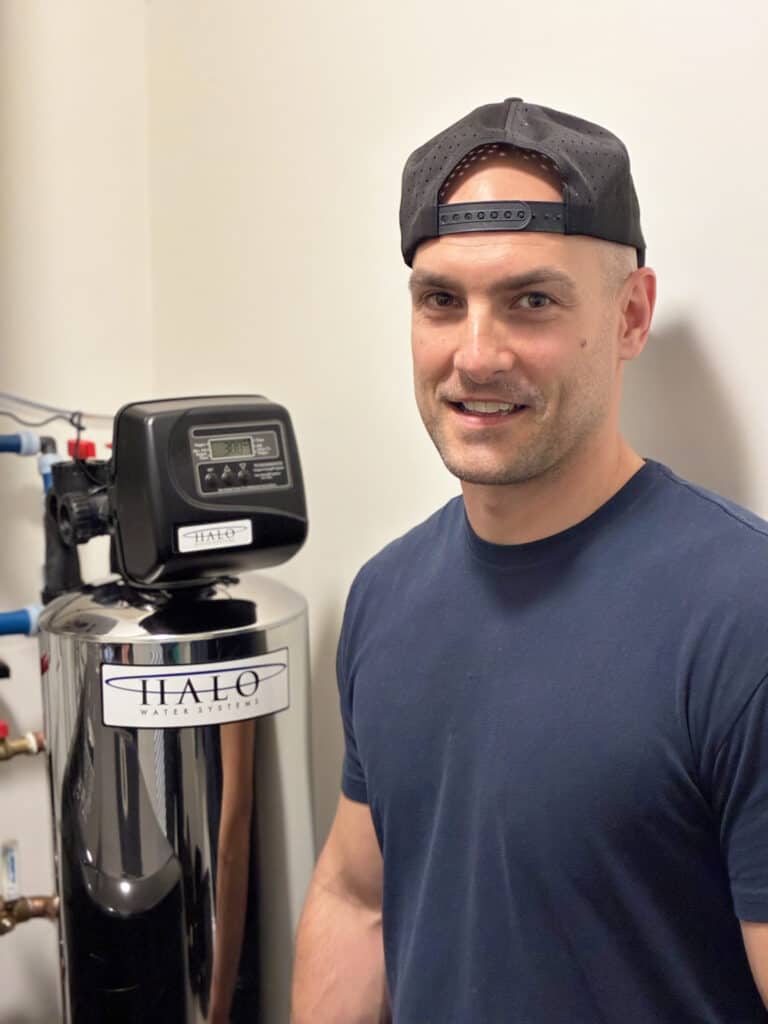 A man in a black cap and navy shirt stands next to a Halo water filtration system in a utility room.