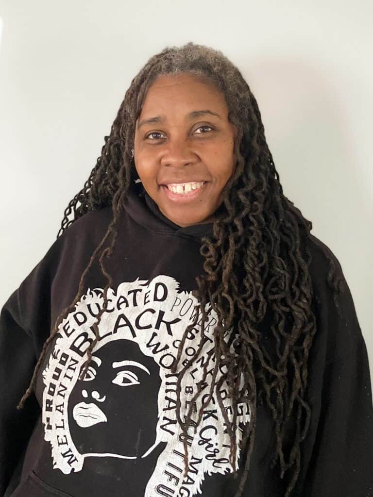 A woman with long, dark, graying dreadlocks smiles at the camera. She wears a black hoodie featuring a graphic of a face and empowering words like "Black," "Educated," and "Proud.