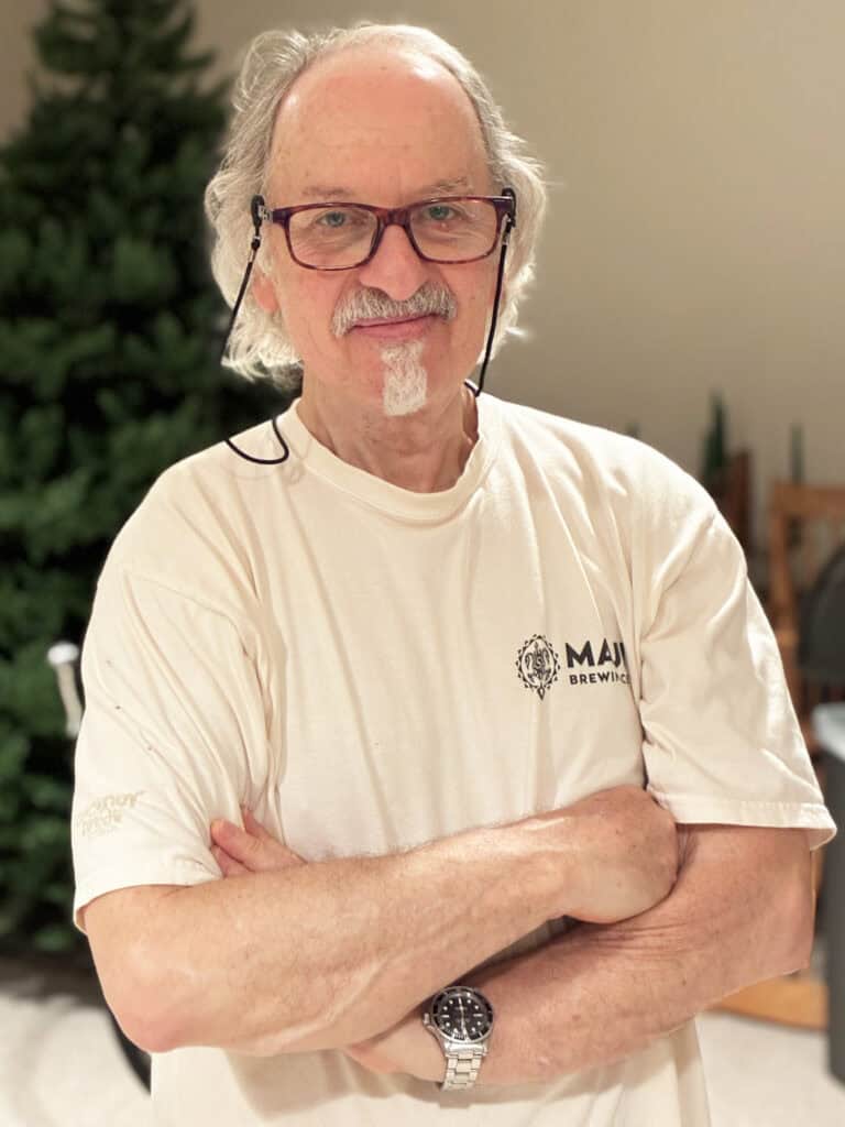 Older man with gray hair and glasses stands indoors with arms crossed, wearing a white “Maui Brewing Co.” t-shirt and a watch. A green tree is visible in the background.